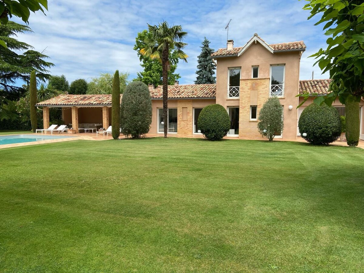 A spacious house is observed, featuring a classic exterior with a tiled roof and multiple windows. Lush green lawns surround the home, complemented by mature trees and neatly trimmed bushes. A covered outdoor area is visible nearby, adding to the tranquil setting.