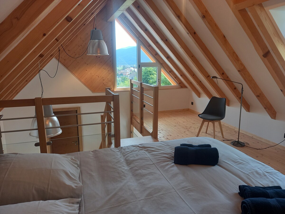 An open mezzanine sleeping area is highlighted by wooden beams and large windows offering views of the Vosges mountains. A spacious bed is positioned prominently, accompanied by neatly arranged towels. A minimalist chair and lamp complement the tranquil space.