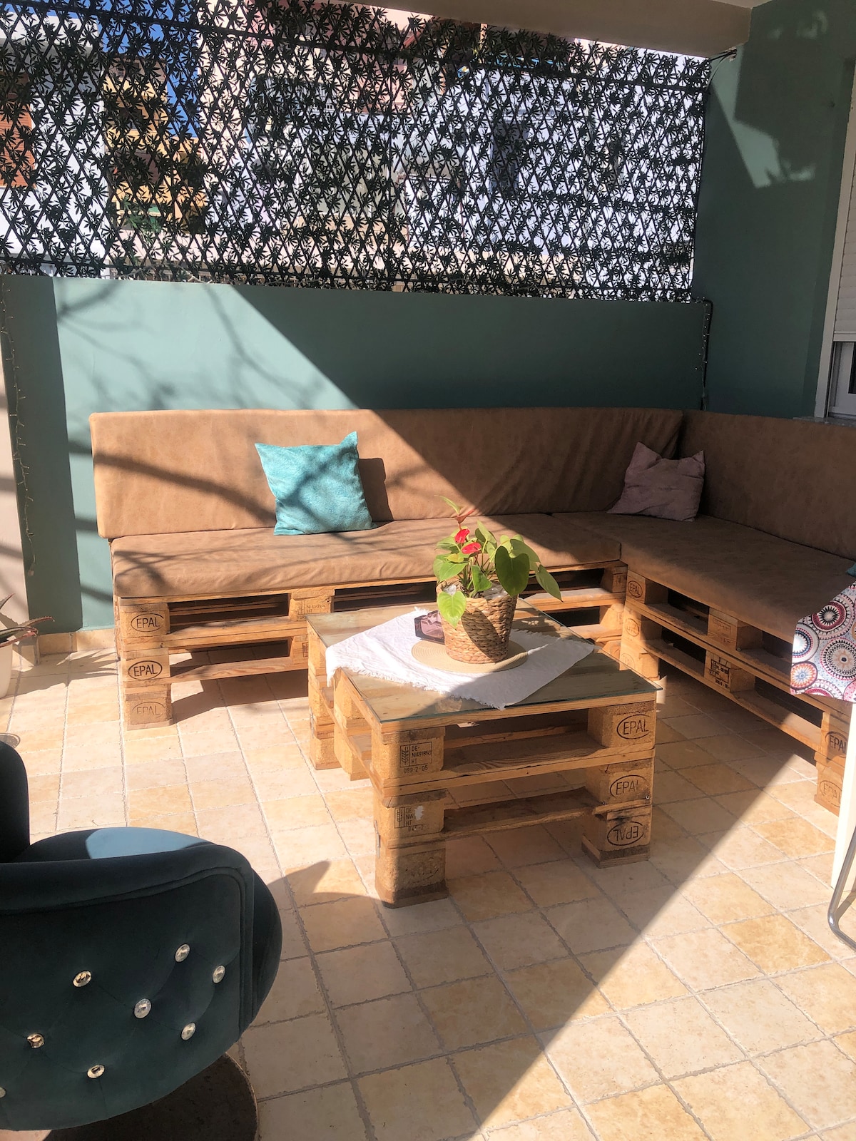 A cozy outdoor seating area is showcased, featuring a sectional couch made from wooden pallets, accompanied by a small table. A potted plant is placed on the table, and colorful cushions add comfort. Sunlight filters through a patterned screen, creating playful shadows.