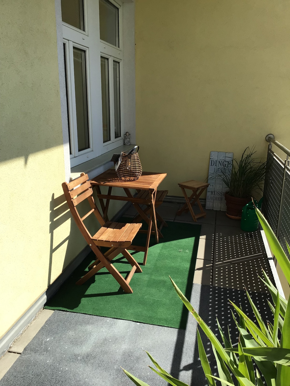 A private terrace is featured with wooden furniture, including a small table and two chairs. Green artificial grass complements the space, which is partially shaded by a railing. Potted plants and decorative items are arranged along the wall, enhancing the area’s cozy appearance.