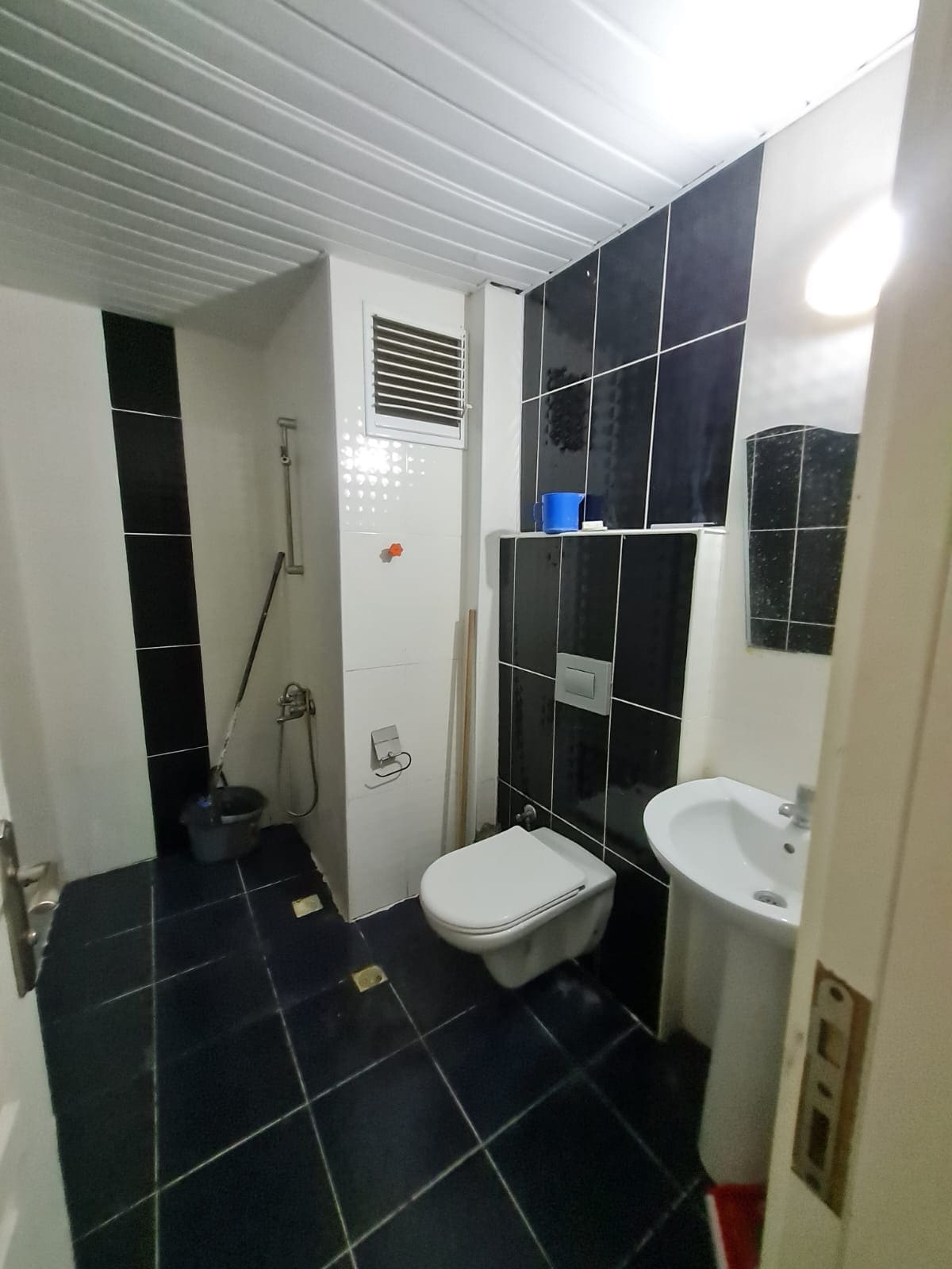 A modern bathroom is presented, featuring a black and white tile design. A toilet and a washbasin are visible, accompanied by a shower area. The space contains minimal furnishings and includes a small window for natural light.
