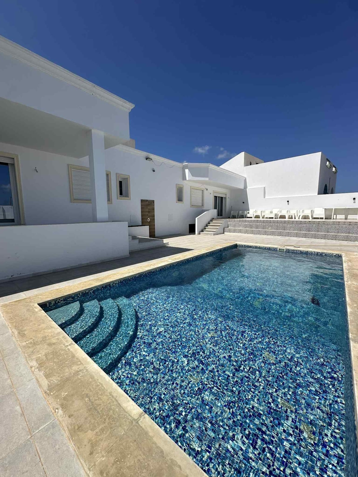 A clear blue swimming pool is visible, inviting relaxation and enjoyment. Surrounding the pool, steps lead to a spacious area featuring white walls and a bright sky above. Several loungers are arranged along the poolside, creating an ideal setting for leisure.