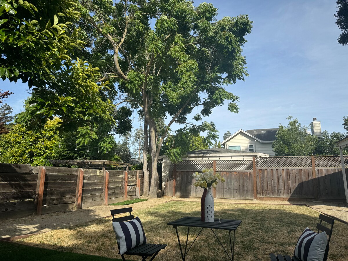 A sunlit backyard is presented, featuring an open grassy area surrounded by wooden fencing. Two black and white striped chairs are arranged around a small table, while a large tree offers shade and greenery in the background.