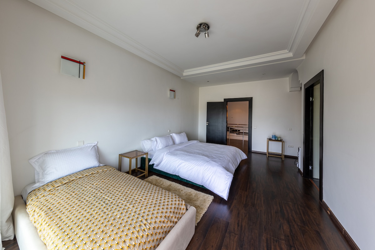 A spacious bedroom features two large beds arranged with white linens, providing a neat and airy feel. A soft rug lies beneath the beds, and a small side table is positioned next to a bed. The room is illuminated by natural light streaming in from a nearby window.