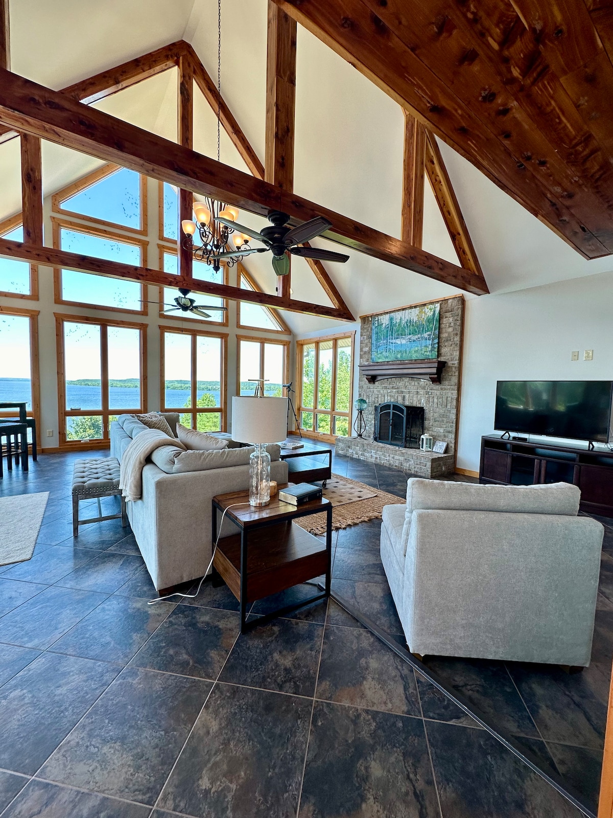 A spacious living area features high ceilings with exposed wooden beams. Large windows provide a view of the lake, while comfortable seating arrangements include a light grey sofa and armchairs. A rustic stone fireplace is positioned below a flat-screen television, adding to the room's charm.