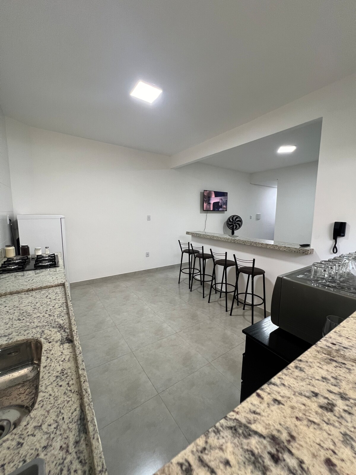 A modern kitchen area features a granite counter and stainless-steel sink. Barstools are positioned at an elevated counter, providing seating for four. A television mounted on the wall adds convenience, while the open layout allows for easy movement between the kitchen and dining space.