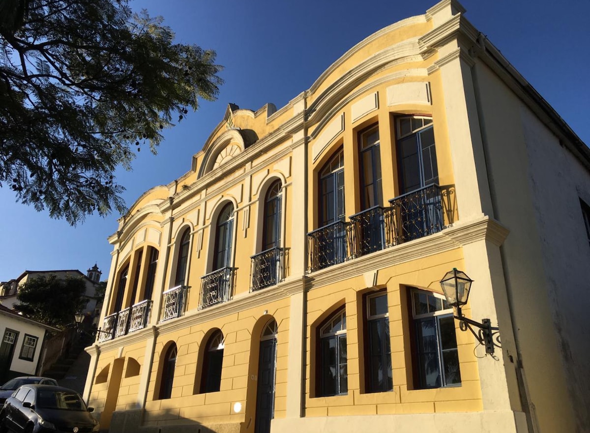 A historic building features a prominent yellow façade with arched windows. Intricate wrought iron railings adorn the windows, while decorative moldings frame the structure. A vintage street lamp is positioned at the side, enhancing the building's charm in the clear blue sky.