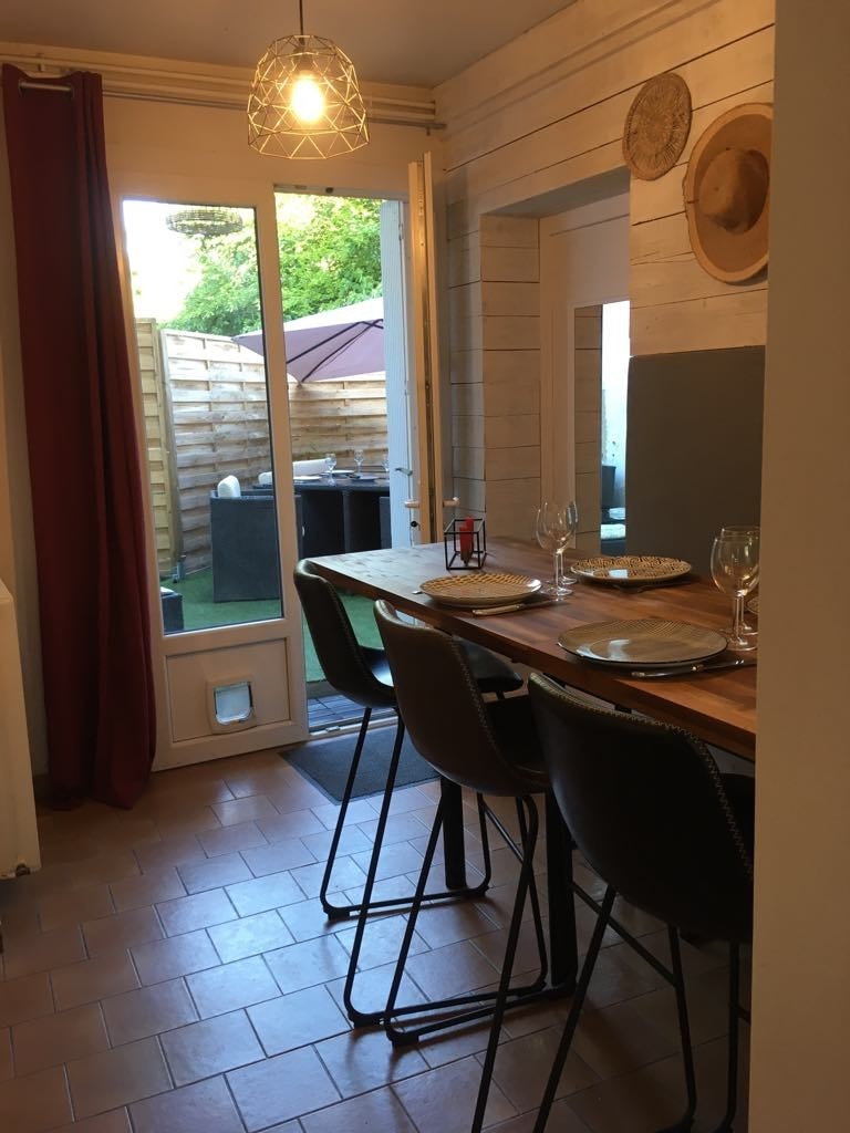 A dining area features a wooden table ready for six guests, set with plates and glassware. Natural light floods through the glass doors leading to a small outdoor space. Soft lighting is provided by a pendant lamp, enhancing the inviting ambiance.