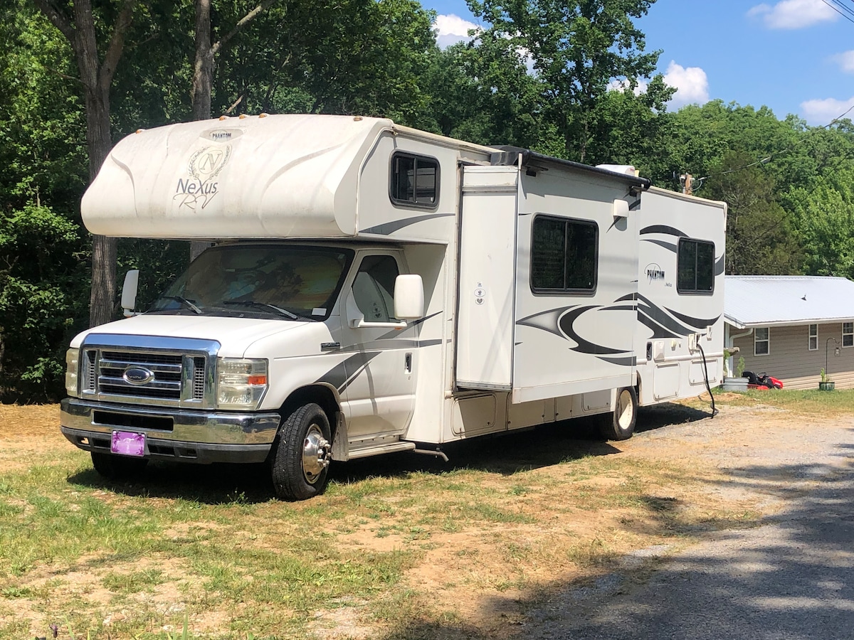 A well-maintained motorhome is parked on a grassy area, surrounded by trees and open sky. The vehicle features a spacious exterior with ample windows and a large awning, providing an inviting space for outdoor relaxation.