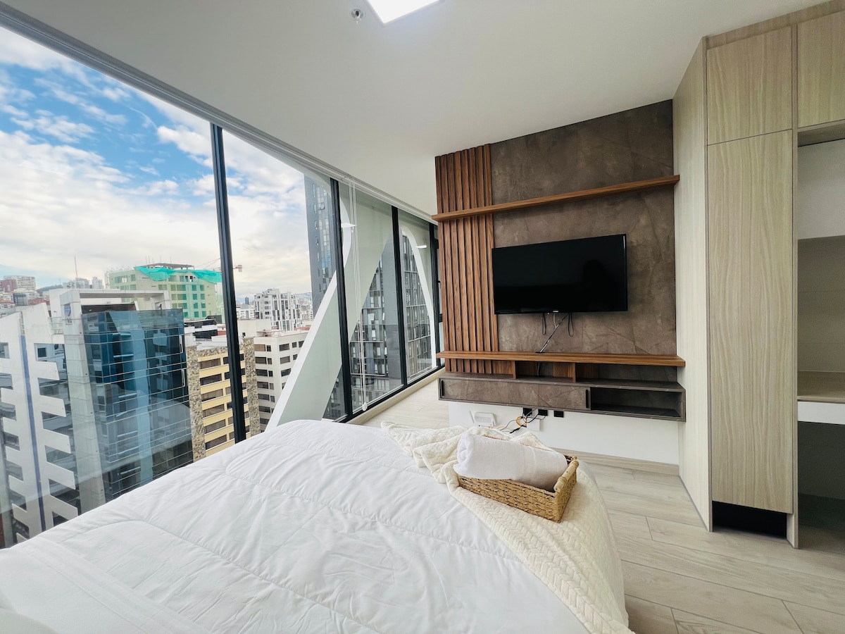 A modern bedroom features a spacious bed dressed in white linens, with a large window providing a panoramic view of the city skyline. A wall-mounted TV is placed above a floating shelf. Natural light fills the room, highlighting the clean lines and neutral tones.