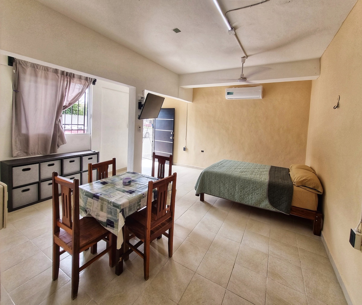 The spacious room features a double bed with light bedding and a decorative throw. A dining table with four wooden chairs is positioned nearby. Natural light enters through a window covered by light curtains, and a neutral color palette enhances the sense of openness.