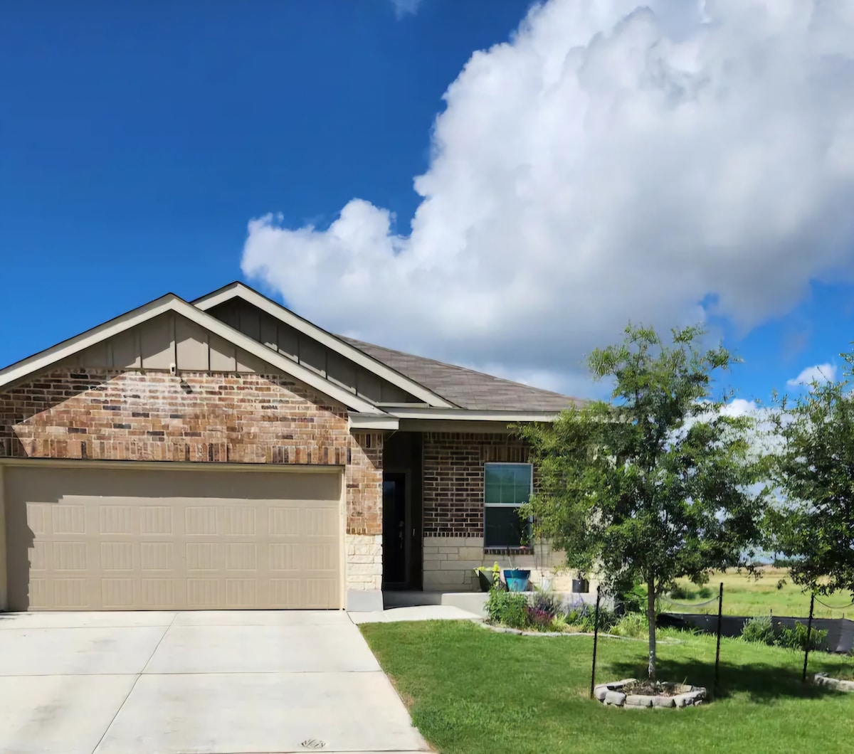 The exterior of the home features a light brick façade with a two-car garage and a concrete driveway. Lush greenery surrounds the entrance, while a clear blue sky and fluffy clouds form the backdrop, creating a peaceful country setting.