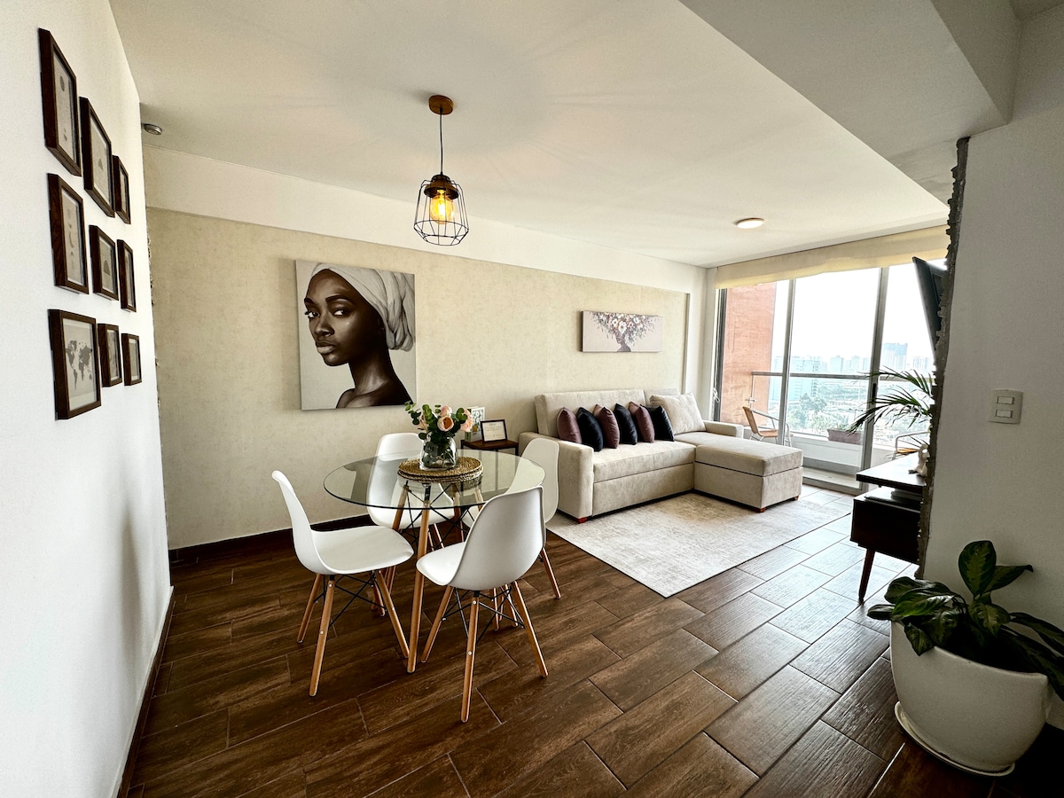A modern living area is highlighted by a comfortable sofa and a glass dining table surrounded by four white chairs. Large windows provide natural light and showcase city views. Minimalist wall art and a potted plant contribute to the chic, welcoming ambiance.