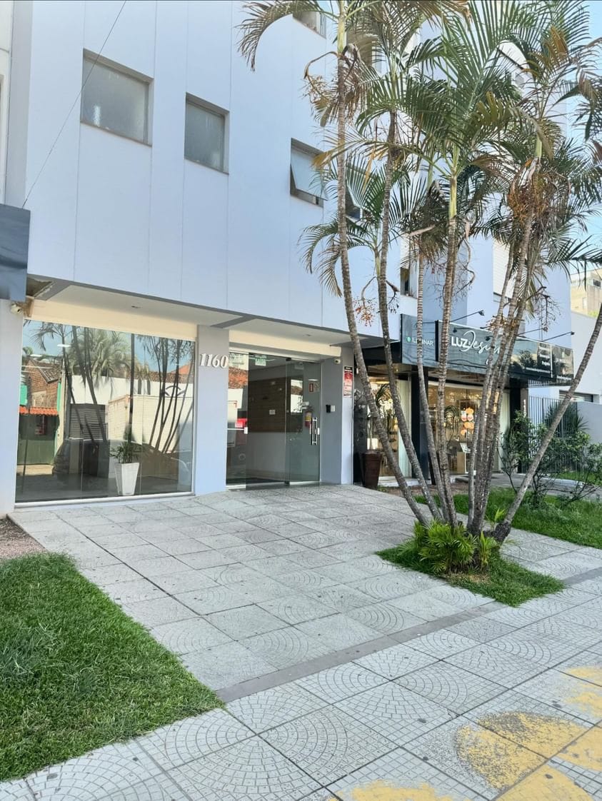 The building's exterior features a modern design with large glass windows reflecting the surroundings. Palm trees line the entrance, and a tiled pathway leads up to a glass door. A small garden area adds greenery to the entrance space.