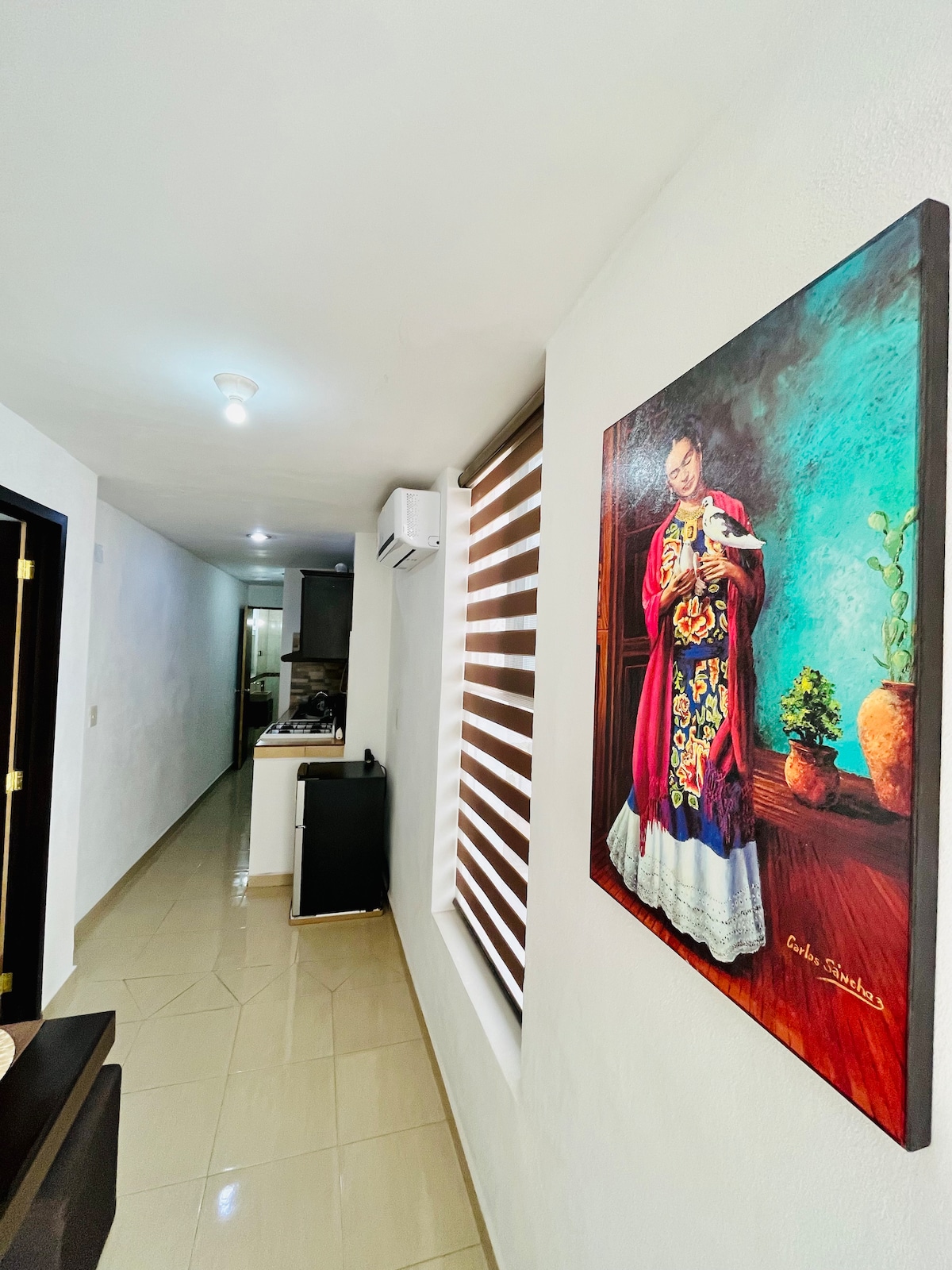 A hallway is depicted, featuring smooth, light-colored tiled floors and white walls. A colorful painting of a woman in traditional attire adorns the wall, providing a vibrant focal point. The view extends towards a kitchen area in the background, where appliances and cabinetry are visible.