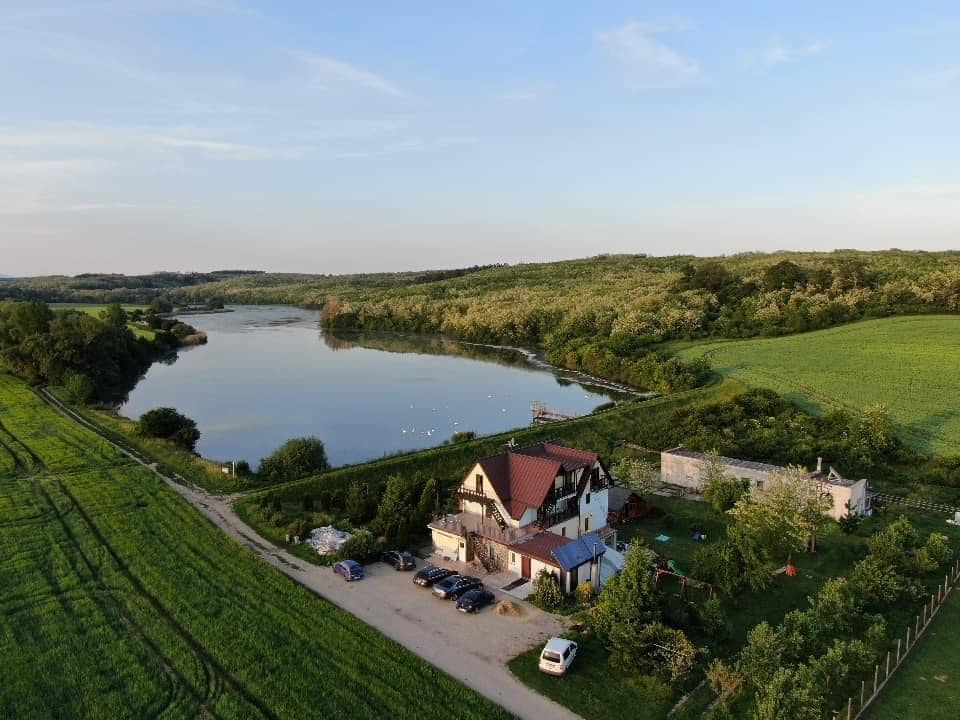 An aerial view captures a serene landscape featuring a lake surrounded by greenery. A charming building with a red roof is positioned near the water, complemented by well-maintained gardens and a gravel pathway lined with parked vehicles.