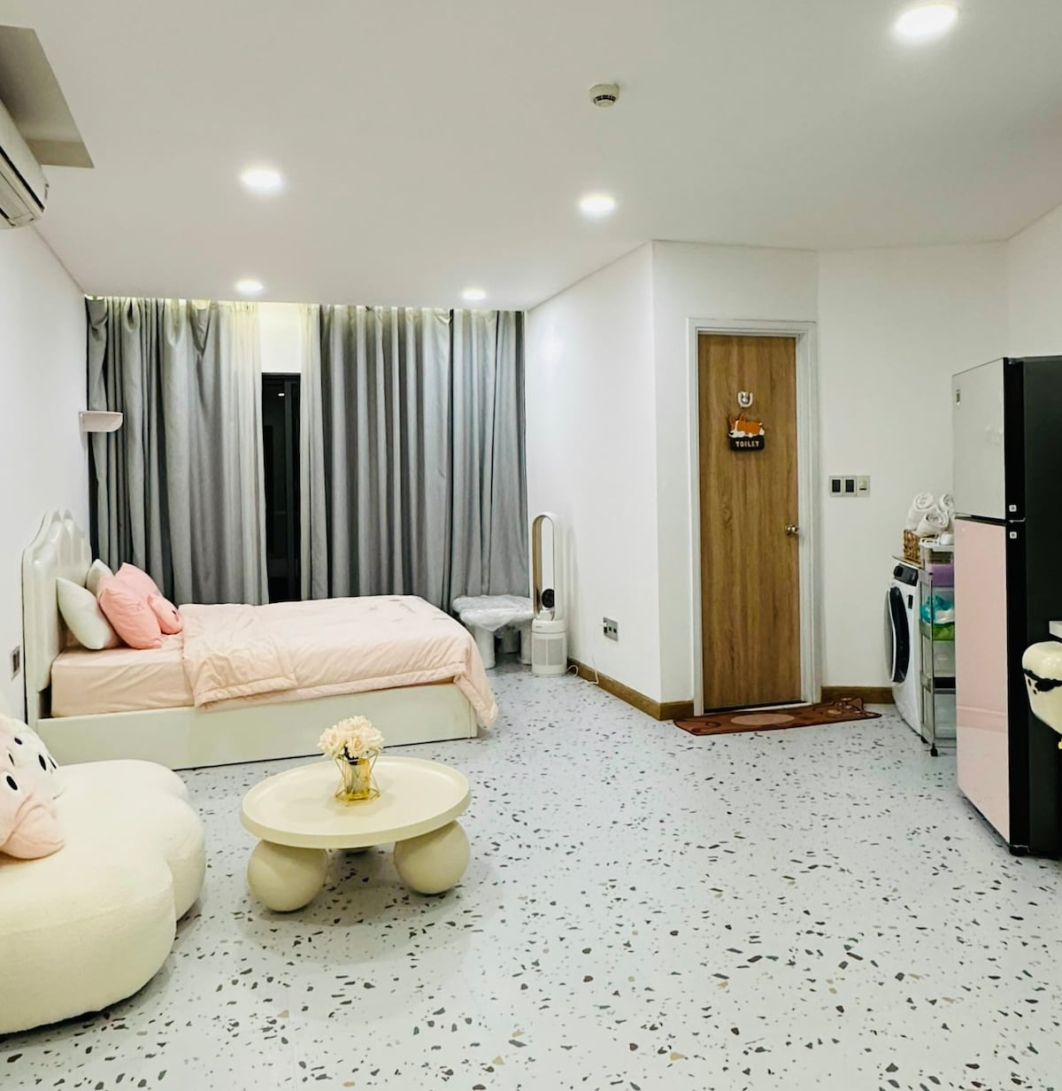 A cozy studio apartment features a light pink bed with soft bedding. A small white table and a plush armchair are positioned across from the bed. Curtains frame a large window, and a door leads to a fridge and washing machine area.