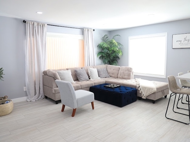 A modern living room features a spacious sectional sofa in light grey, complemented by a navy ottoman and a cozy throw blanket. Natural light streams through the large windows, with soft sheer curtains providing a gentle backdrop to the harmonious decor.