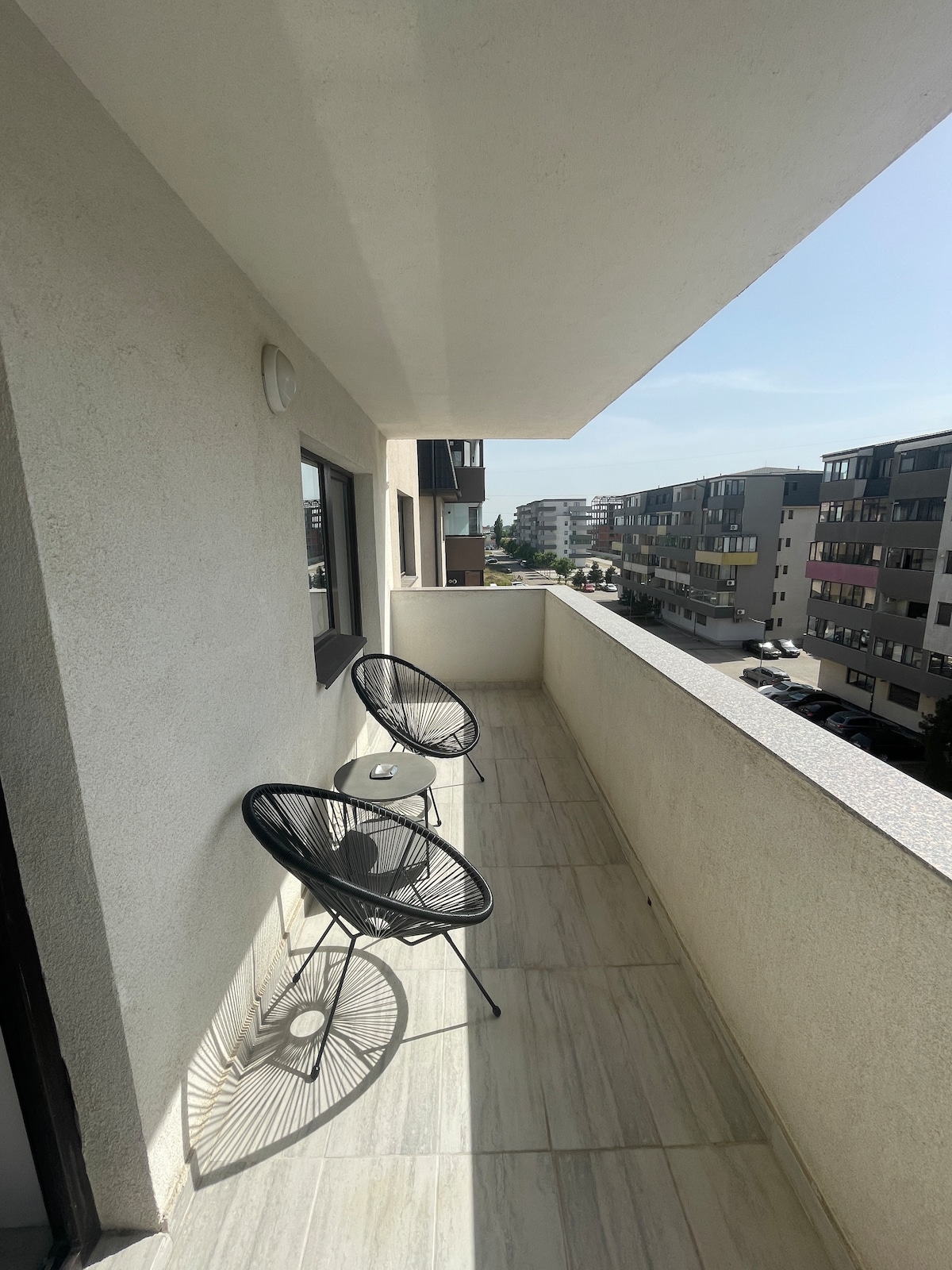 A narrow balcony is featured, furnished with two round black chairs and a small table. The floor is tiled with light marble, and a clear view of surrounding residential buildings is visible beyond the balcony railing.