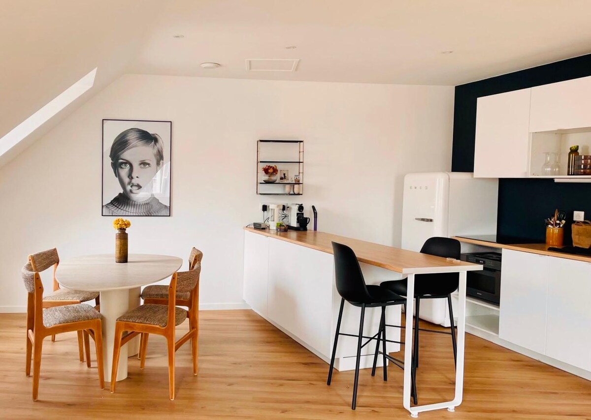 An airy kitchen and dining area features a round table with four chairs and a minimalist design. A sleek bar counter complements the space, while modern appliances and a decorative shelf create an organized and inviting environment. Natural light floods in through the angled ceiling.