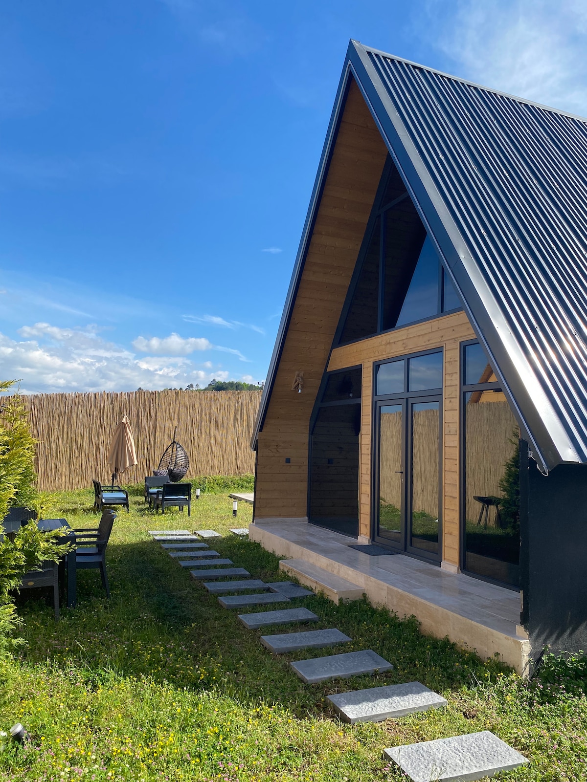A modern A-frame bungalow is set against a clear blue sky. The front entrance features large glass doors, and a path of stepping stones leads to the beautifully landscaped yard, which includes outdoor seating areas and a gentle border of greenery.