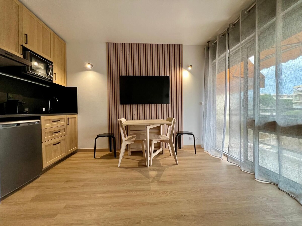 An open kitchen area is designed with wooden cabinetry and a stainless steel refrigerator. A round dining table is surrounded by four simple chairs. A mounted TV is positioned on a striped accent wall, with sheer curtains allowing natural light to filter in.