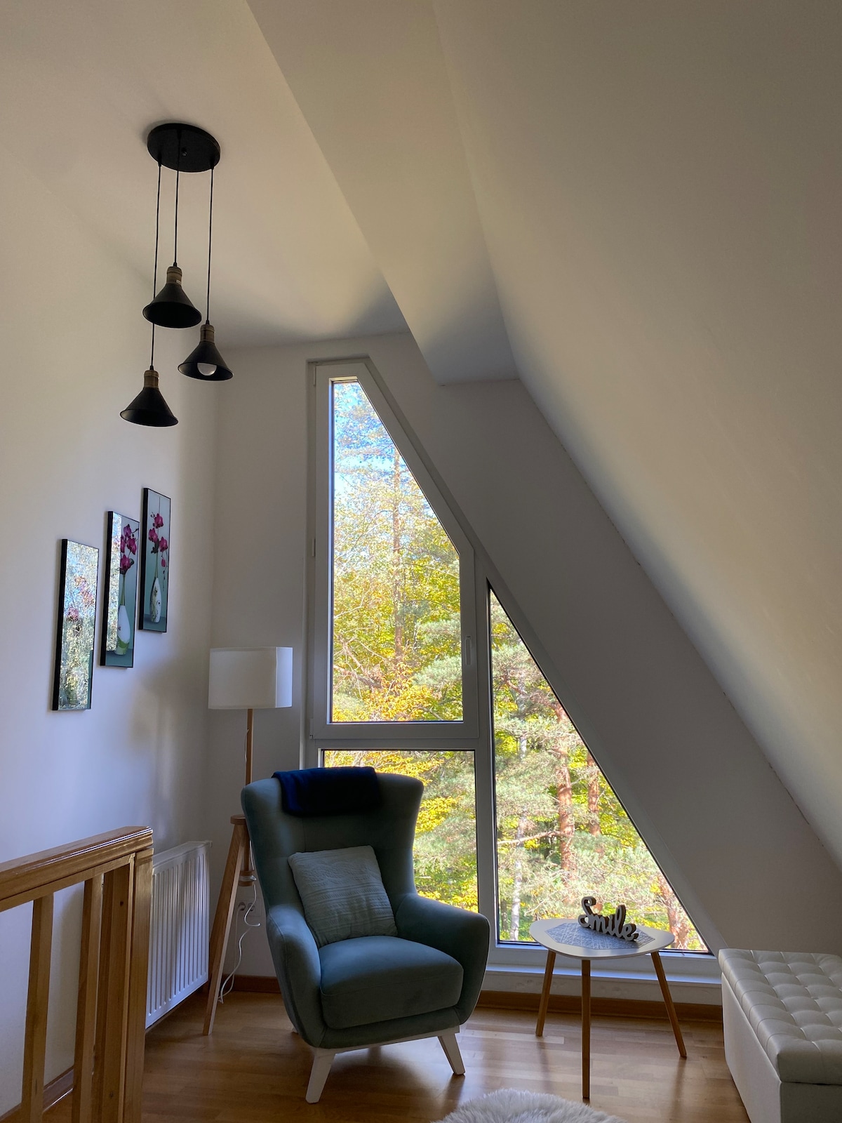 A corner nook features an angled ceiling with three pendant lights. A contemporary green chair is positioned near a large triangular window, offering views of trees outside. A small round table is placed beside the chair, adding to the cozy atmosphere.