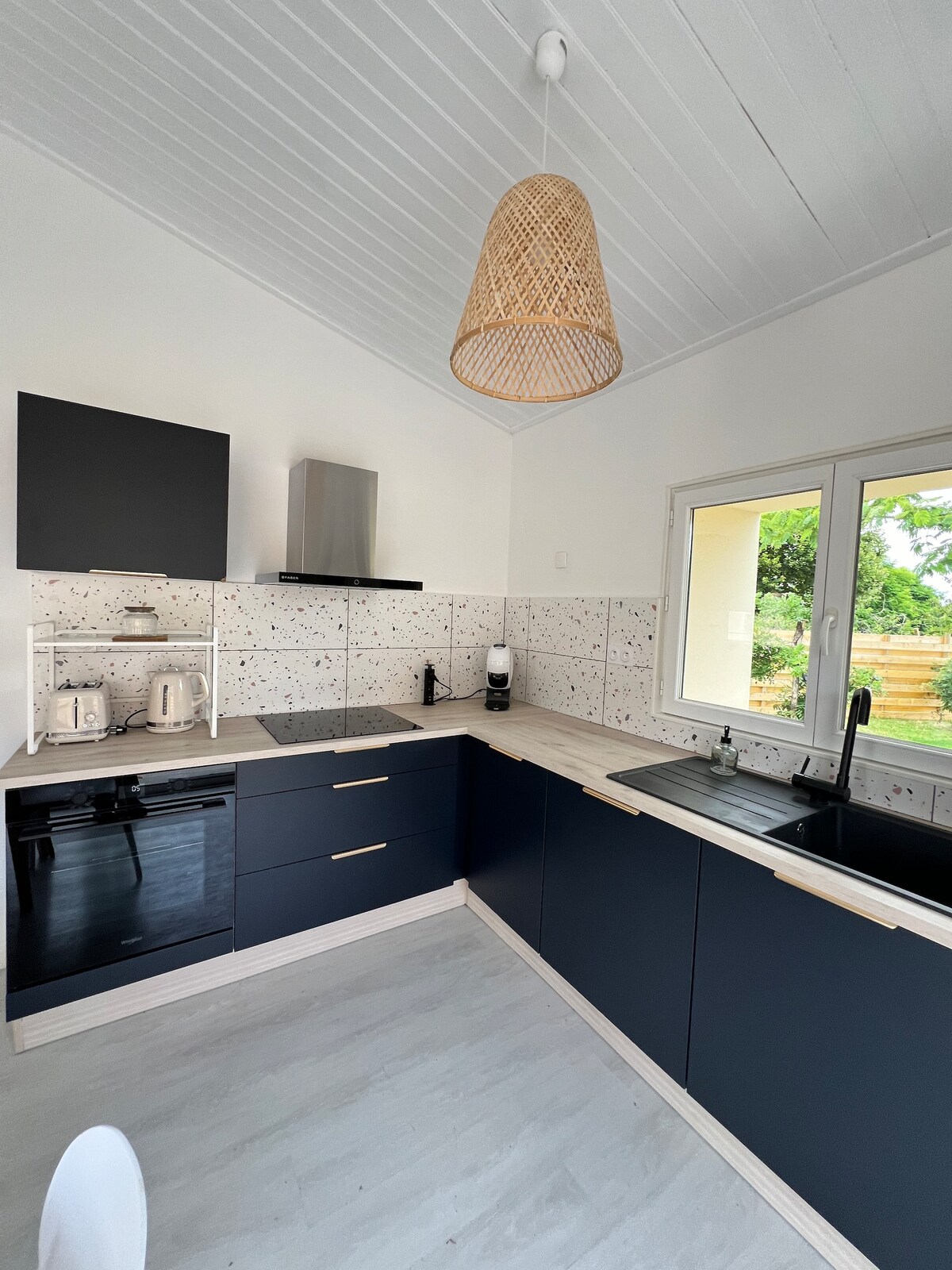 A modern kitchen features sleek black cabinetry paired with light wood accents. A shiny stainless-steel refrigerator and stove are positioned for convenience. Natural light floods in through a large window, showcasing views of the garden. A decorative woven light fixture hangs from the ceiling.