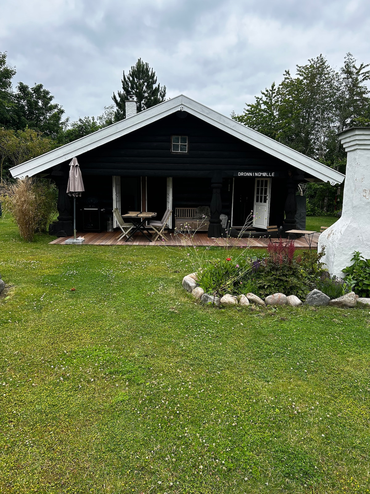 The charming black house features a spacious wooden deck, surrounded by lush greenery. A table and chairs are set up for outdoor dining, while a large umbrella provides shade. Flower beds with various plants enhance the inviting outdoor space.