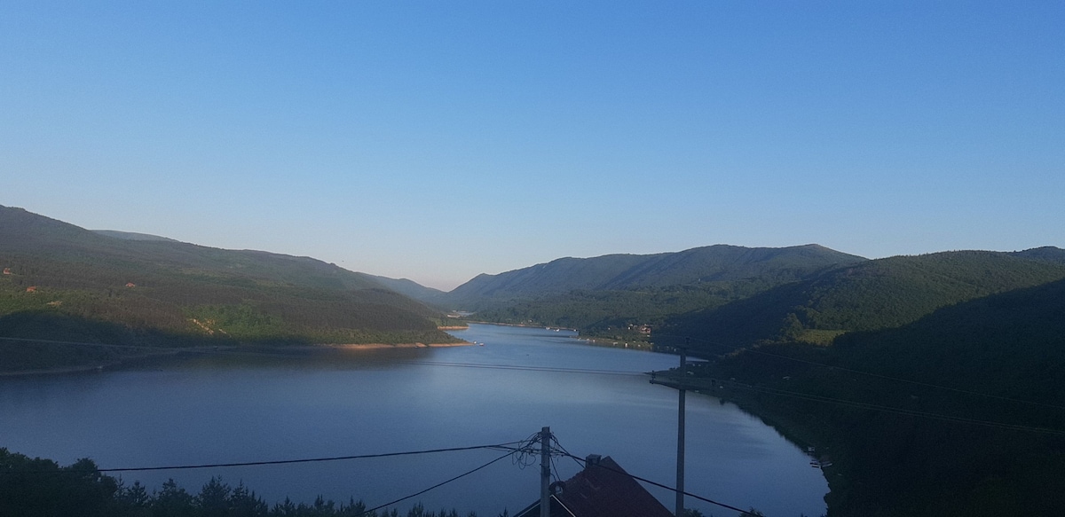 A panoramic view of a serene lake is captured, bordered by lush green mountains under a clear blue sky. The calm water reflects the surrounding landscape, creating a tranquil atmosphere. Power lines are visible in the foreground, leading to the distant hills.
