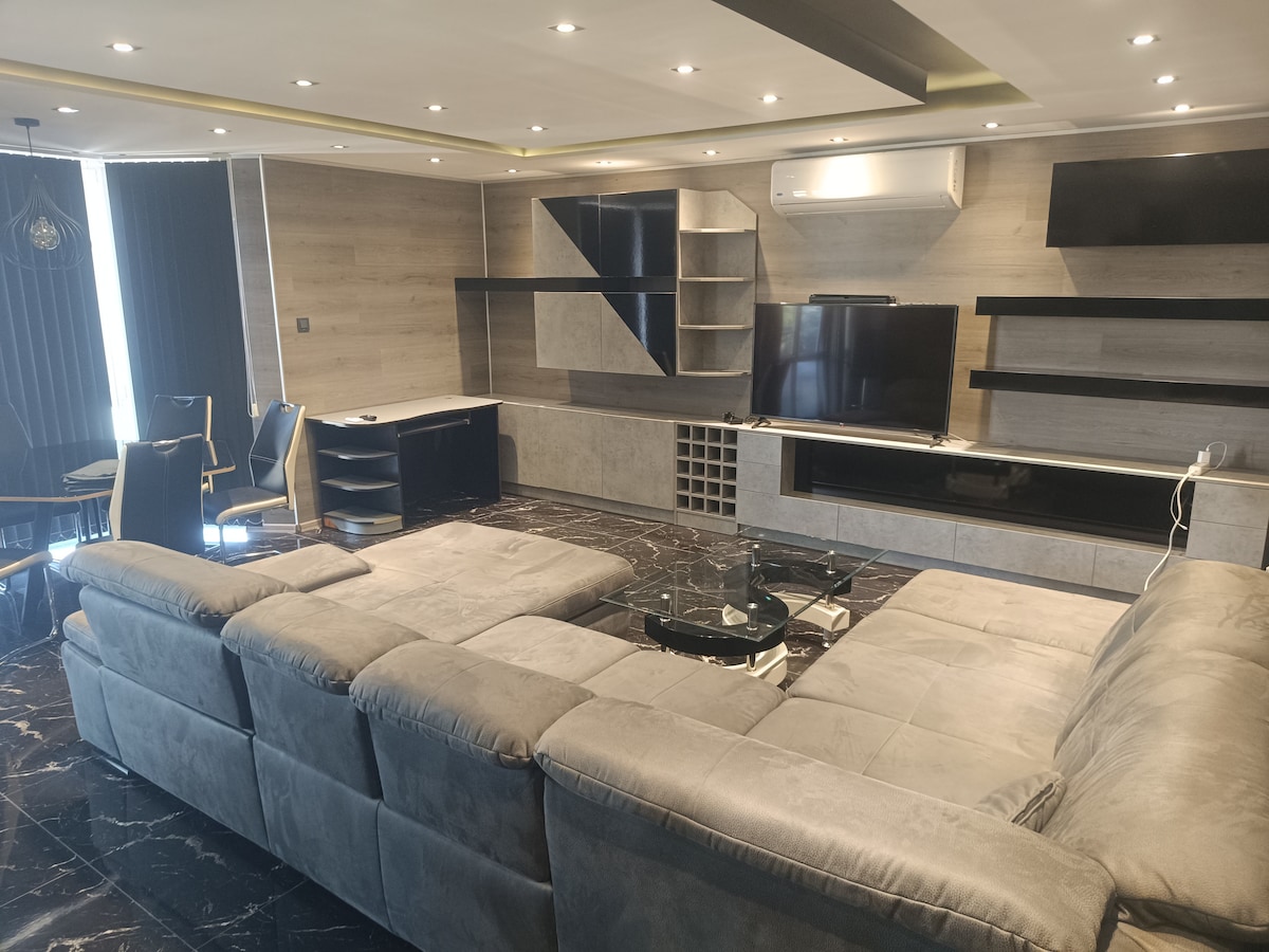 A spacious living room is featured with a large sectional sofa positioned around a glass coffee table. A sleek television is mounted on the wall, accompanied by stylish shelving units. Natural light filters through dark curtains, enhancing the marble flooring and modern design.