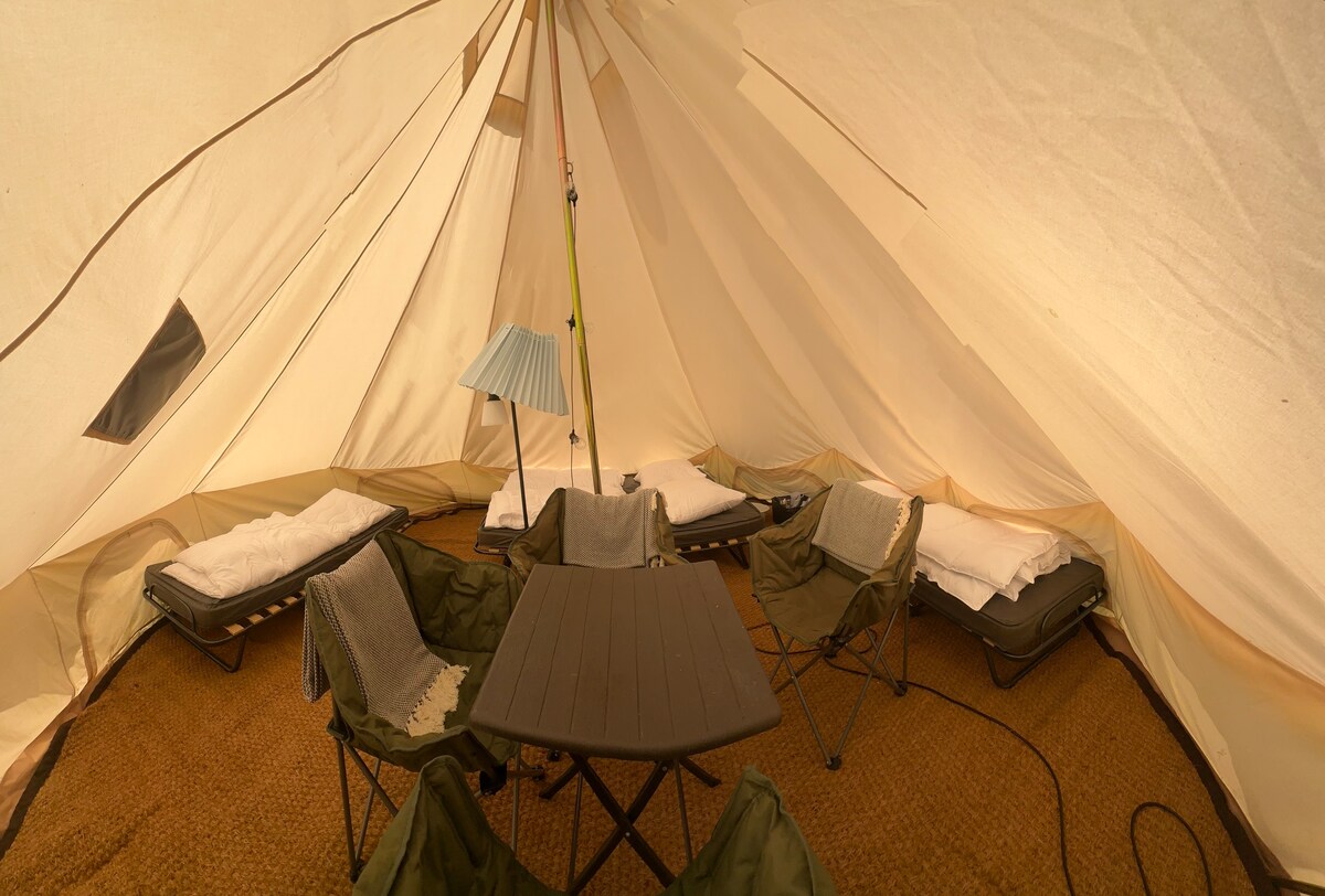 Inside the tent, several foldable cots are arranged, each equipped with pillows and duvets. A small rectangular table occupies the center of the space, while a lamp provides illumination. The tent's fabric ceiling is high, and natural light filters through the small windows.