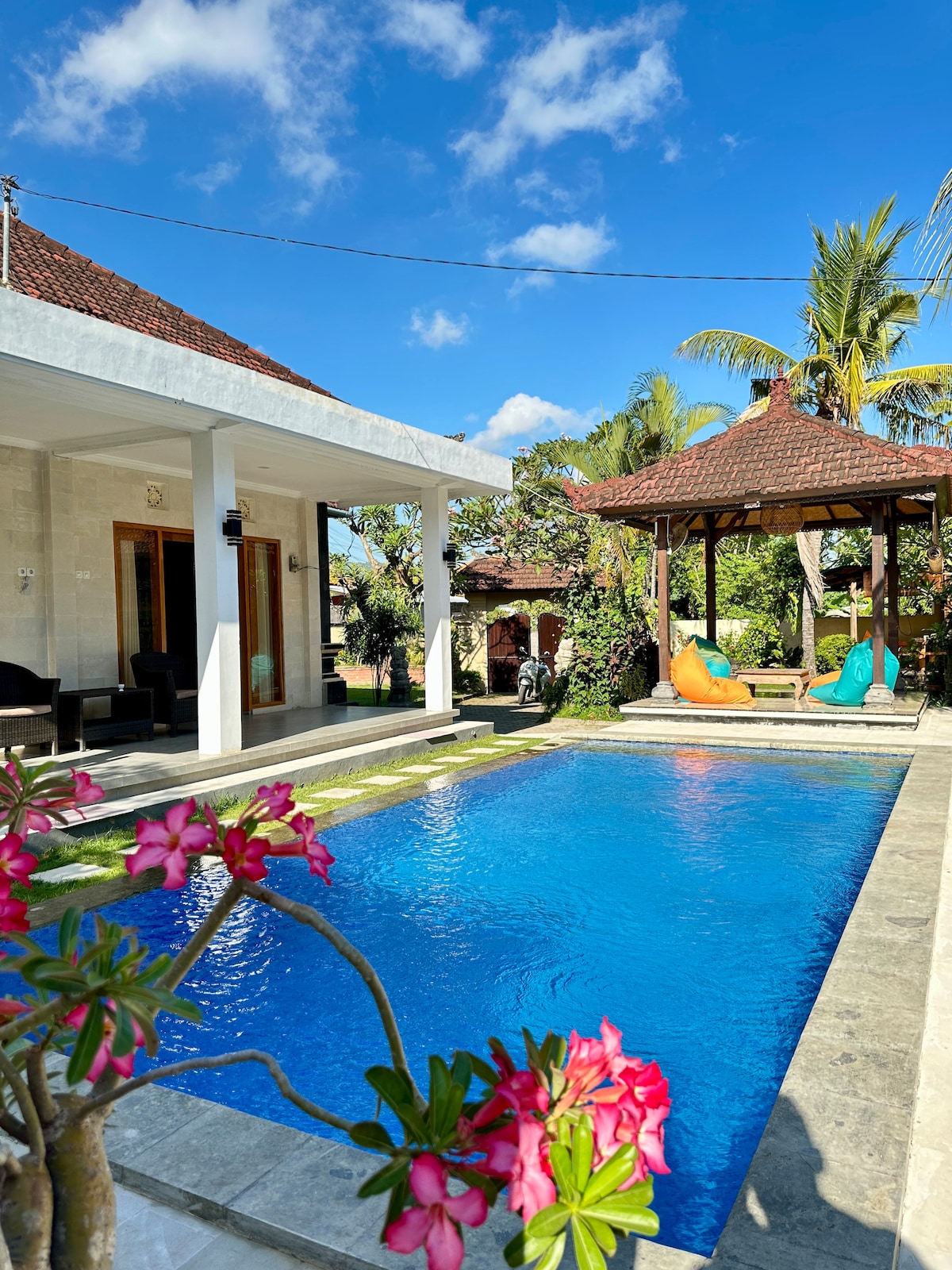 A serene outdoor space showcases a clear blue swimming pool surrounded by lush gardens. A traditional gazebo offers shaded seating, while colorful cushions provide comfort. Nearby, vibrant flowers add natural beauty, complementing the inviting atmosphere of the property.