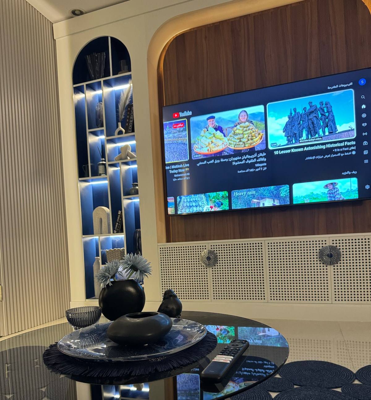 A modern entertainment area features a large flat-screen television mounted on a wooden wall. A round table is set with decorative items, including a dark vase and a floral centerpiece, surrounded by subtle shelves displaying art pieces and ambient lighting.
