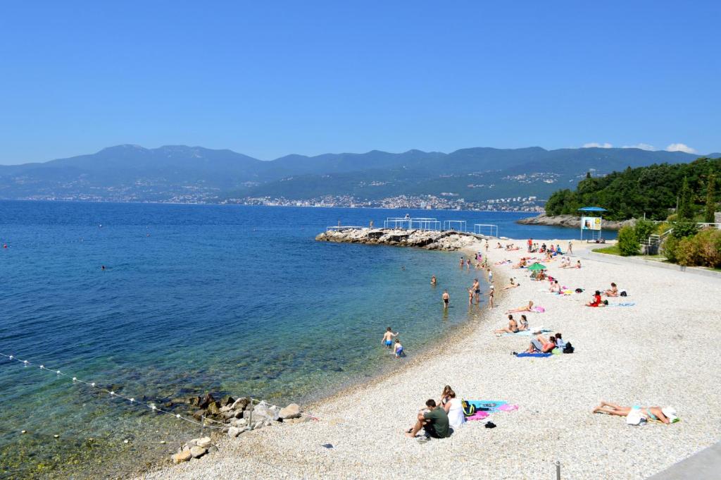 A serene beach scene is presented, featuring a pebbled shoreline meeting clear blue waters. Sunbathers are scattered along the beach, with lush greenery and a mountainous backdrop creating a tranquil ambiance. Umbrellas and loungers may be seen in the distance, emphasizing a relaxed, outdoor experience.
