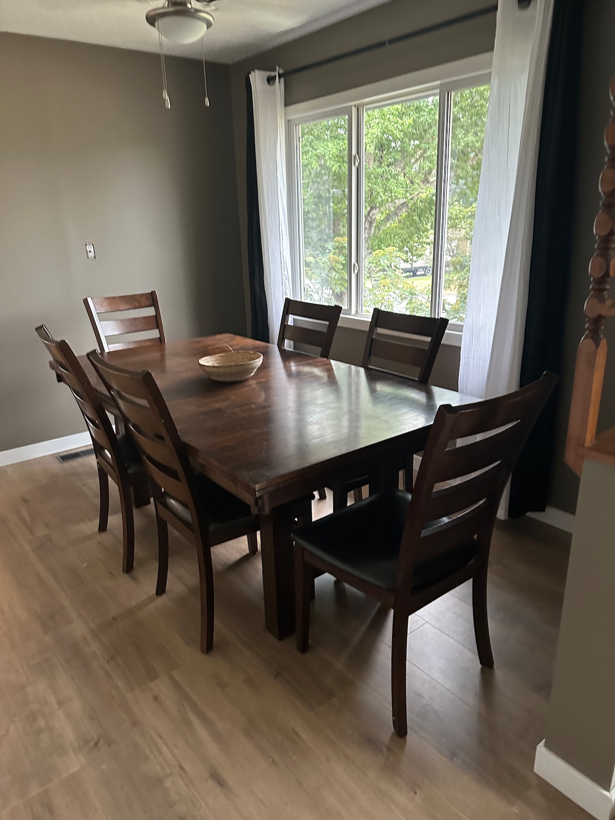 A large wooden dining table is surrounded by six brown wooden chairs with black cushions. Natural light filters through a large window dressed with sheer white curtains, illuminating the room with a calm atmosphere. The floor displays a light wood finish.
