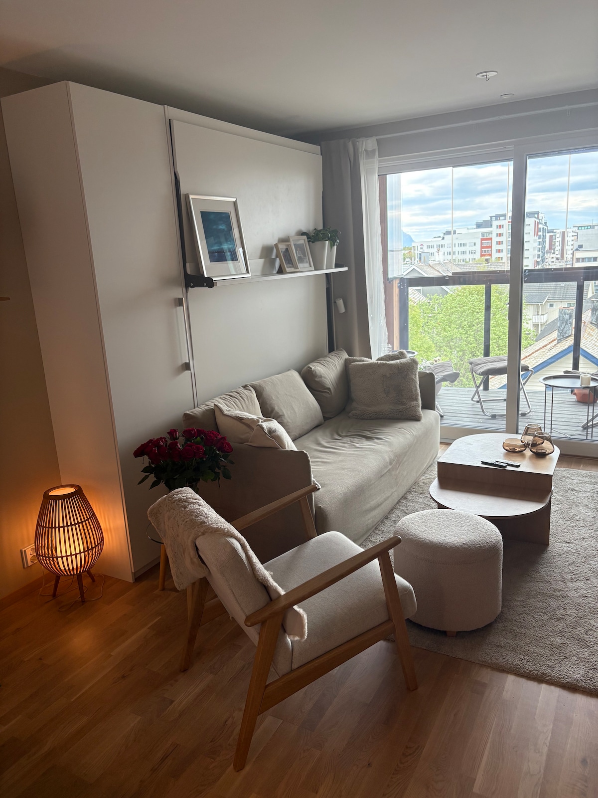 A cozy seating area is featured with a light-colored sofa adorned with soft cushions. A wooden coffee table holds two glasses, beside a small ottoman. A floor lamp provides soft illumination, and large windows offer views of the outside greenery.