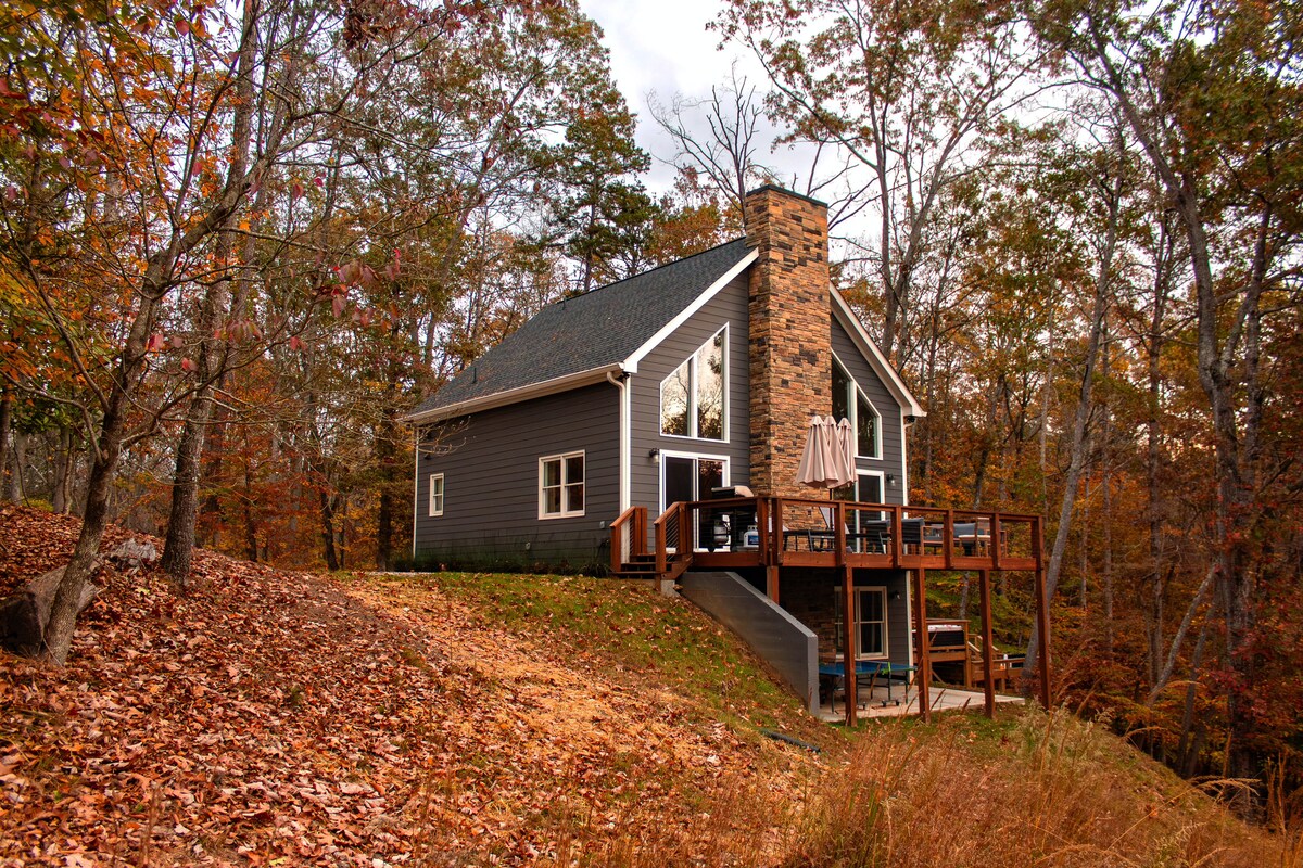 A contemporary home is nestled among autumn trees, showcasing a wooden deck with outdoor seating. A stone chimney rises from the side, and fallen leaves cover the ground, indicating the season. The structure features large windows that allow natural light to enter.
