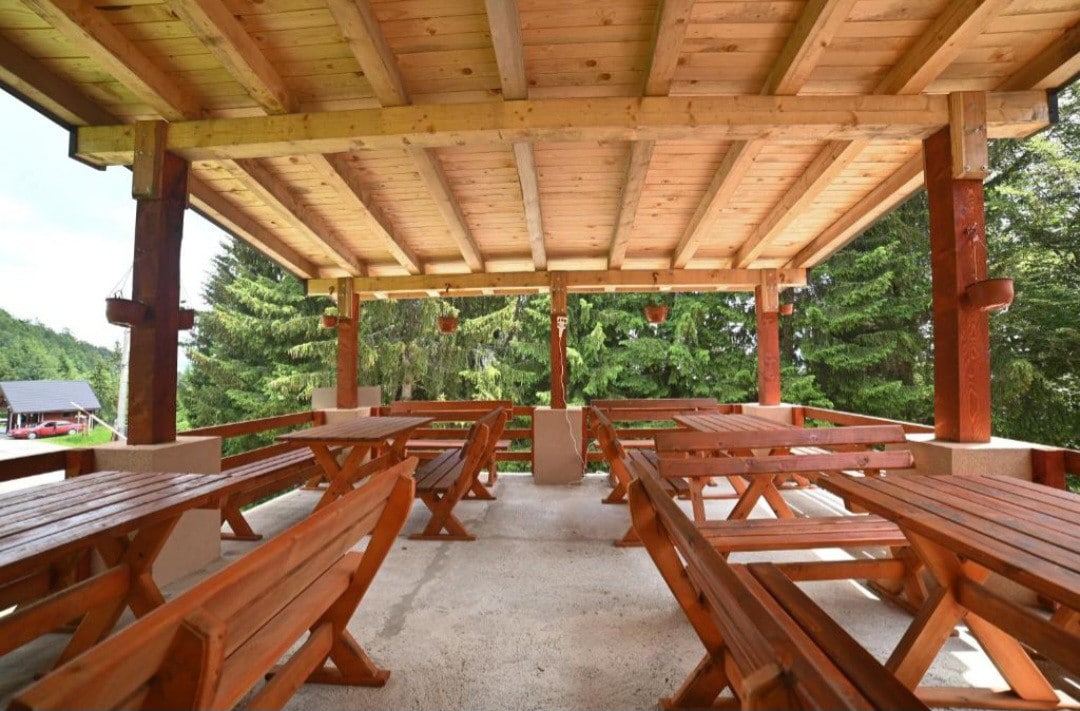 An open-air dining area features sturdy wooden tables and benches arranged under a wooden-beamed roof. Lush greenery can be seen in the background, creating a natural and spacious setting. The concrete floor complements the rustic atmosphere.