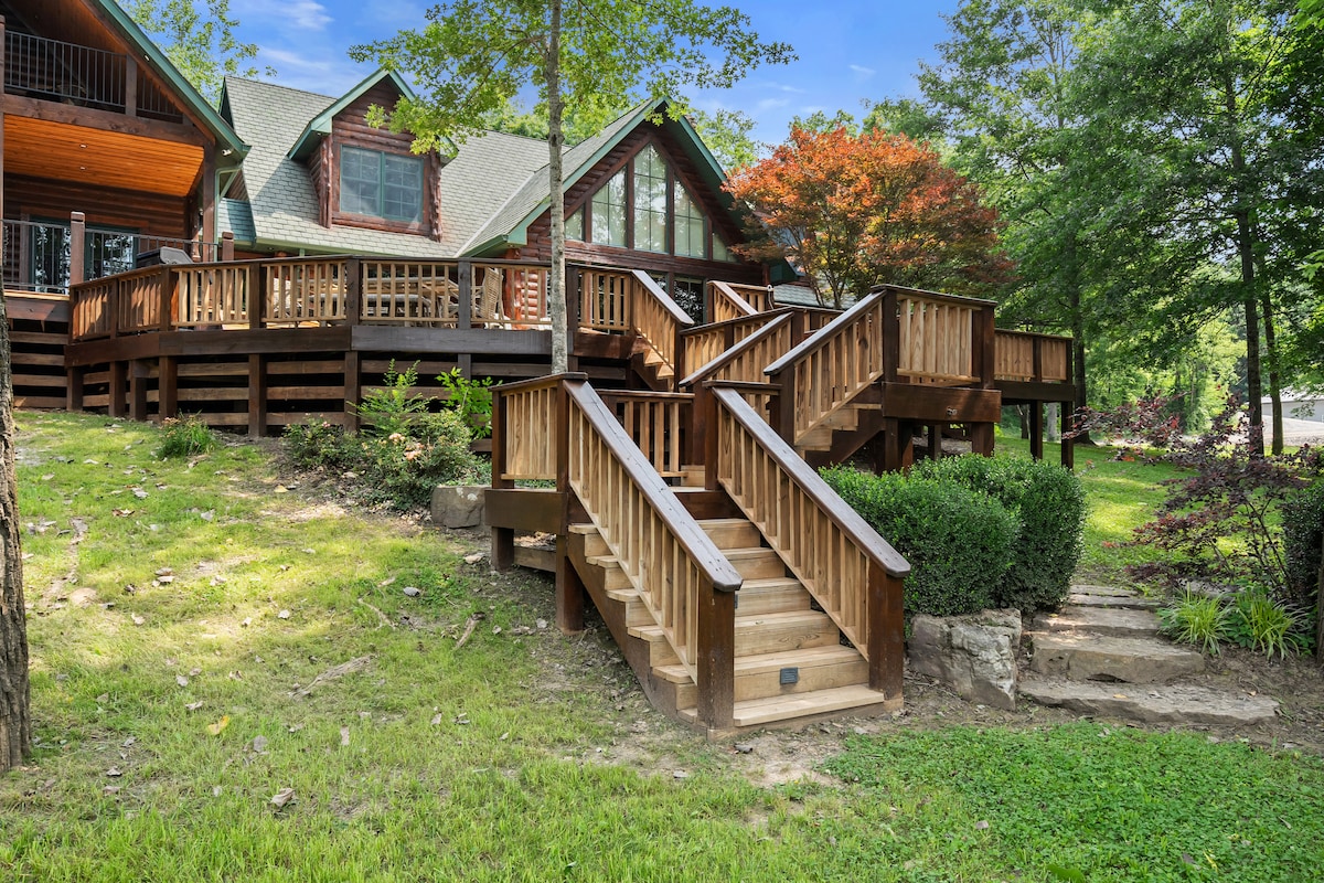 The lodge features a multi-tiered wooden deck with staircases leading to multiple entry points. Surrounding greenery and trees provide a natural setting, enhancing the outdoor spaces available for relaxation and gathering.