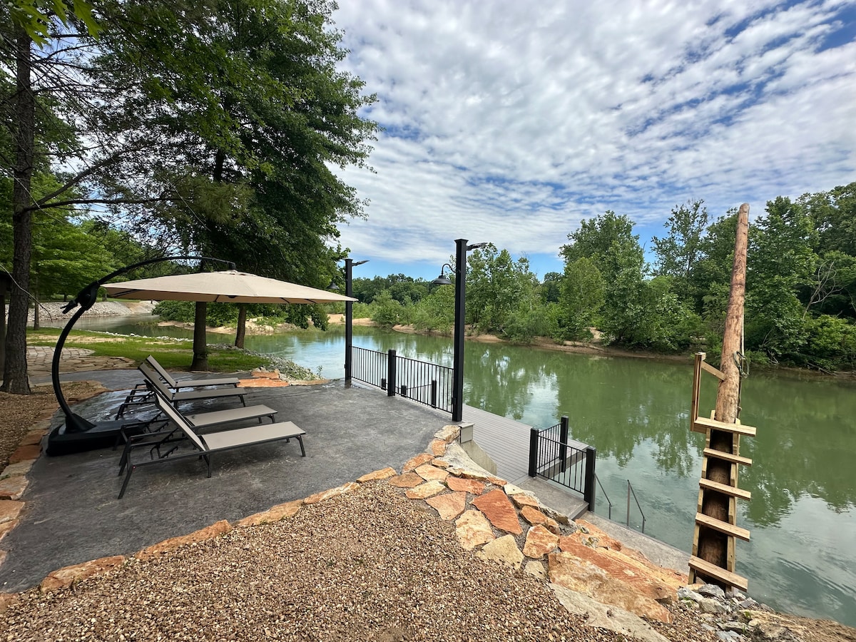 A gravel area leads to the serene Current River, with gentle green waters reflecting the surrounding trees. Lounge chairs provide a place to relax under a large umbrella. A wooden dock is accessible, featuring a ladder for easy entry into the river.