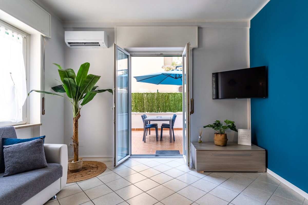 A bright living area features a comfortable sofa and decorative elements, including a potted plant. Large glass doors open to a terrace with outdoor seating under a blue umbrella, while a wall-mounted TV adds to the entertainment options. Natural light enhances the welcoming ambiance.