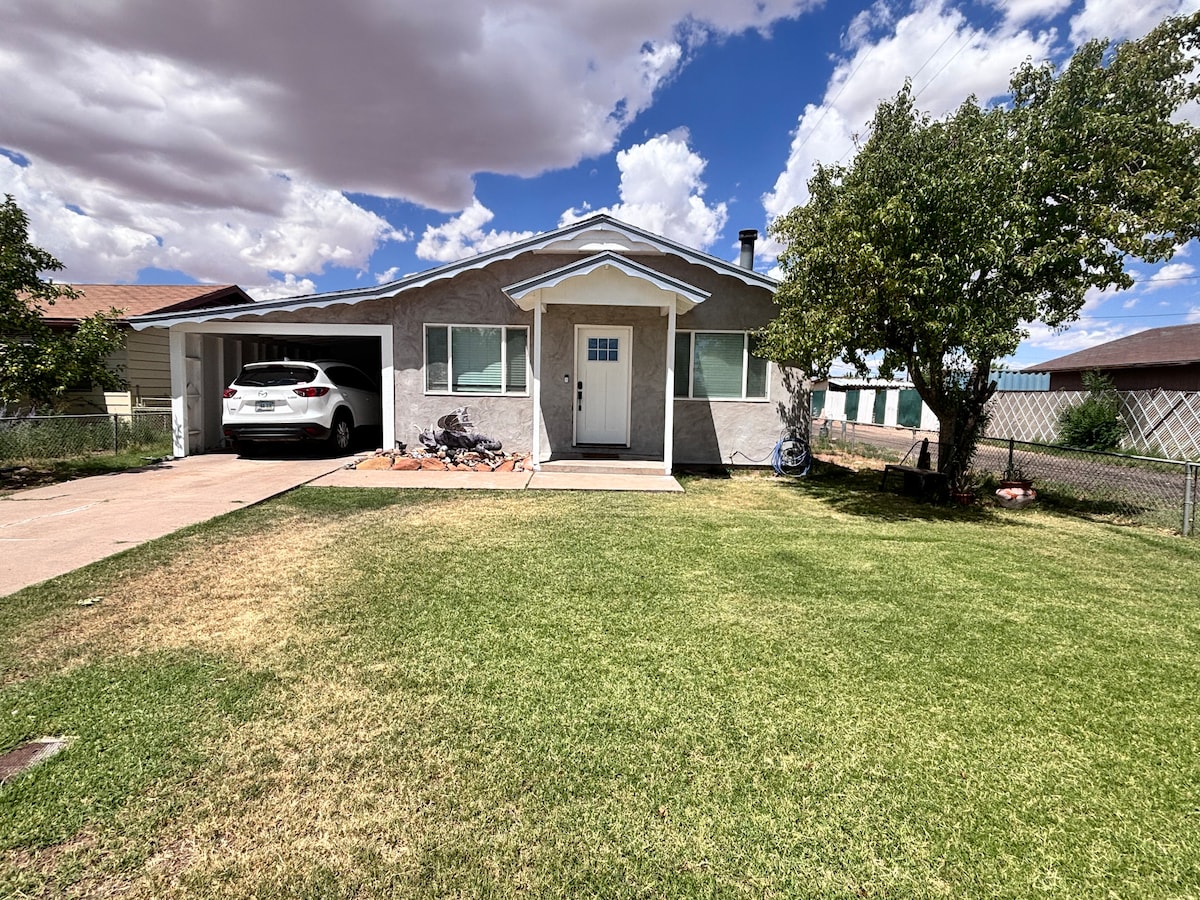 A single-story home is set against a backdrop of partially cloudy skies. The front yard features well-kept grass, and a small porch leads to a central door. A driveway is visible, accommodating a parked vehicle beside a bicycle. Trees provide subtle greenery around the property.