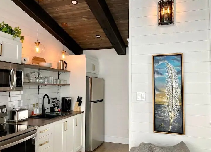 A modern kitchenette features stainless steel appliances and open shelving with glassware. An artistic wall hanging adds visual interest, while warm pendant lights illuminate the space. Exposed wooden beams contribute to the overall design, creating a welcoming atmosphere.