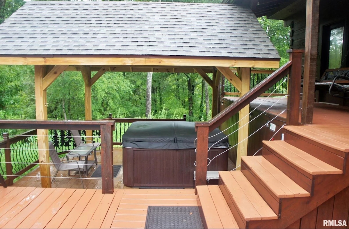 An outdoor deck features a covered hot tub surrounded by wooden railings and steps. Nearby, a small seating area is visible beneath the shelter, with lush greenery in the background adding to the secluded atmosphere.