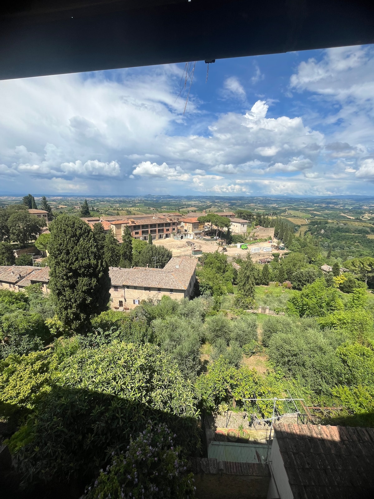 A panoramic view of the surrounding hills and countryside is visible, showcasing lush greenery and the historic buildings of San Gimignano. The sky is partly cloudy, creating a serene backdrop for the tranquil landscape.