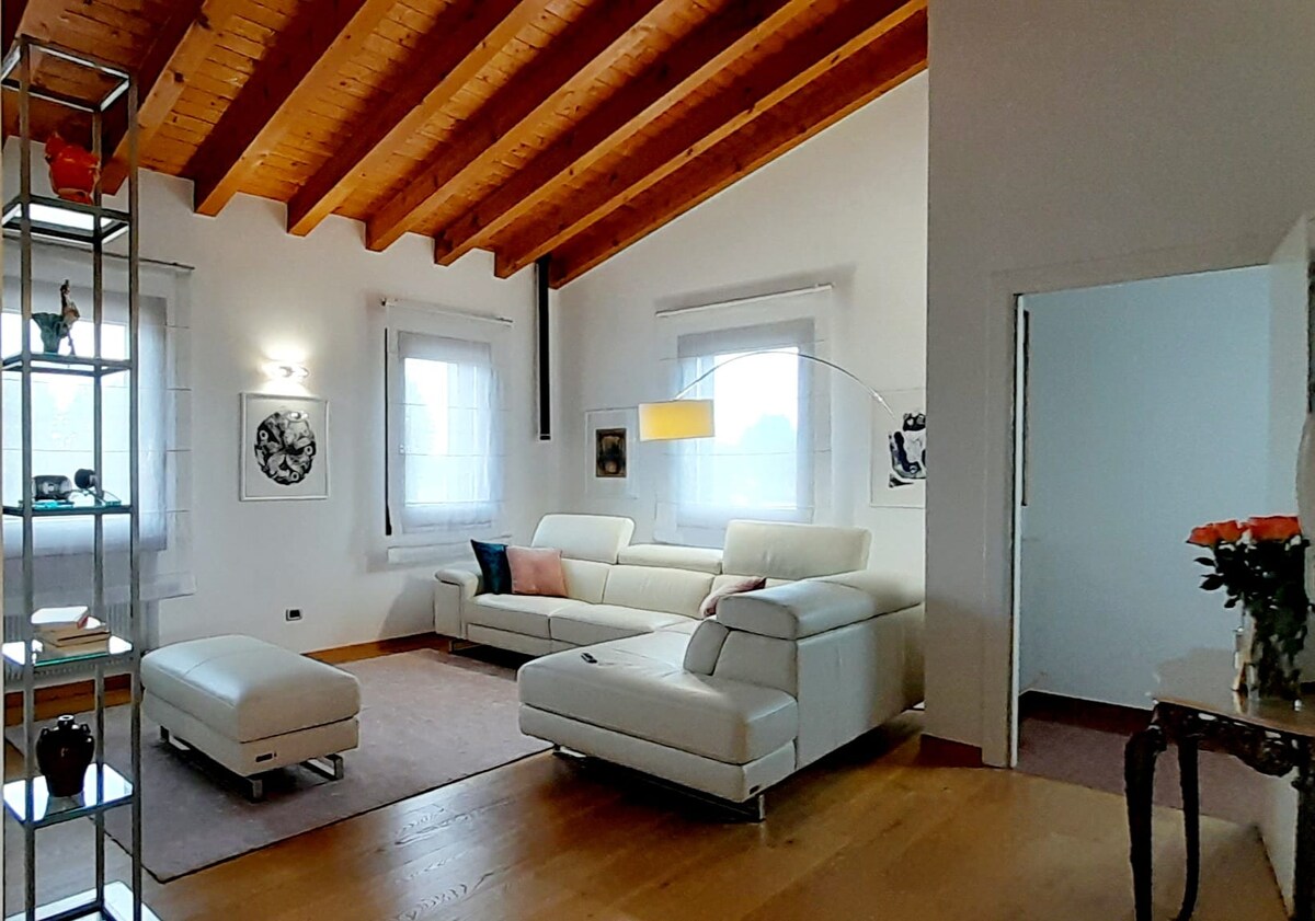 The spacious living area features a white leather sofa and matching ottoman, complemented by wood beams on the ceiling. Natural light enters through large windows adorned with sheer curtains, while a modern floor lamp provides additional illumination. Decorative elements include wall art and a vase of roses.