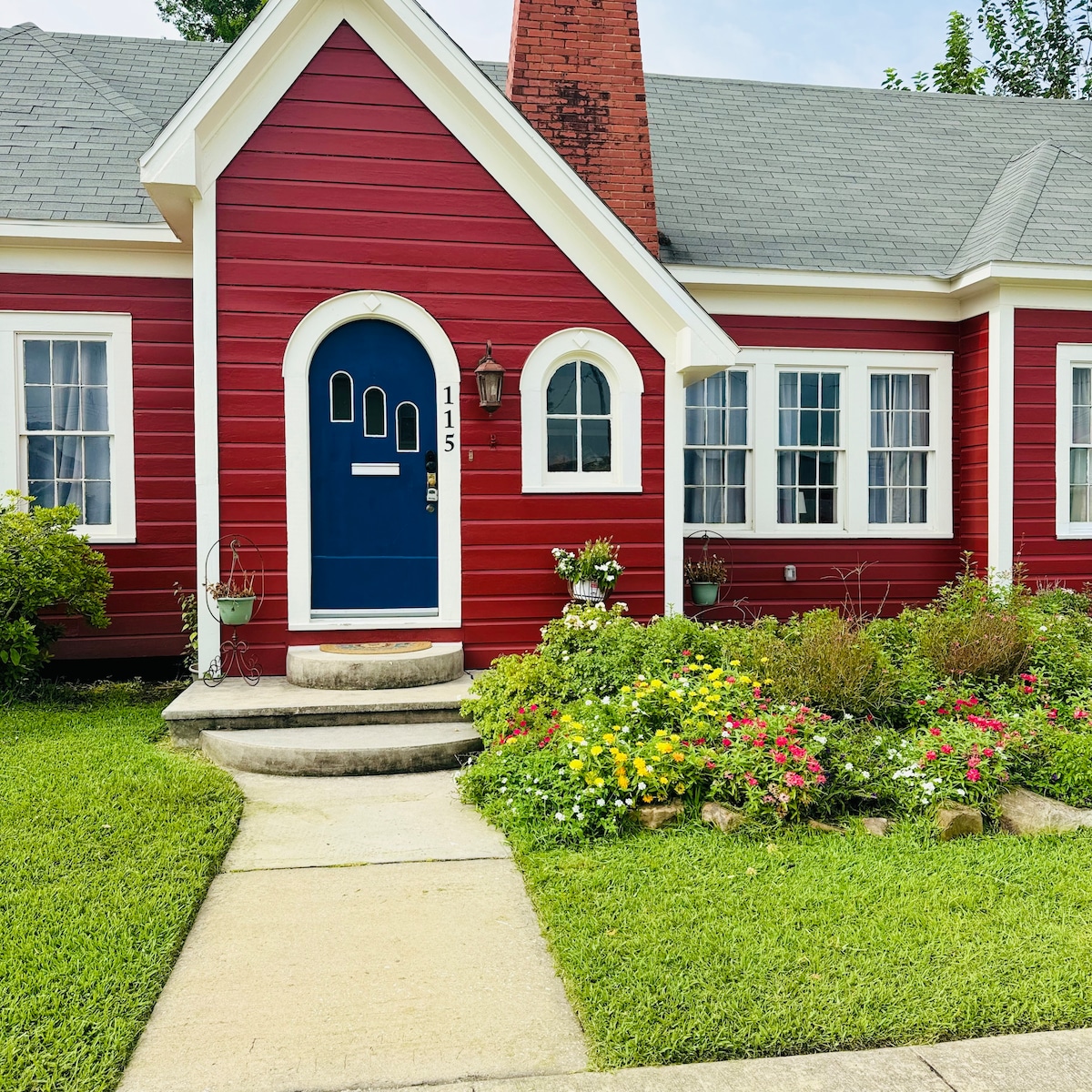 The exterior of a charming red cottage is displayed, featuring a blue front door with a unique window design. Colorful flower beds bloom alongside a neatly cut lawn, and a stone pathway leads to the front steps, complemented by decorative plants in hanging pots.