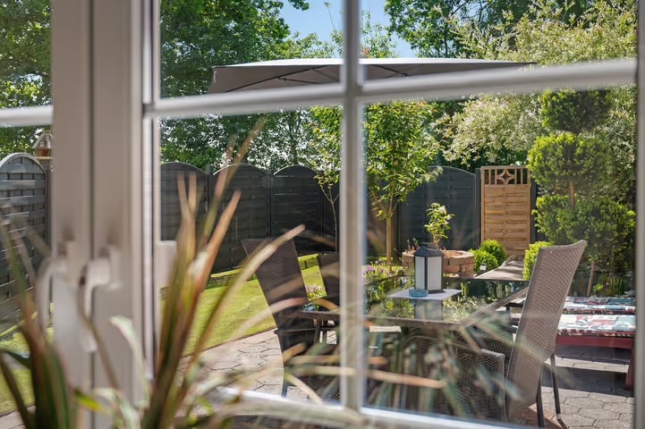 A view through a window frames a serene garden, featuring a table with chairs and an umbrella. Lush greenery surrounds the space, while a small wooden structure is visible in the background, offering an inviting atmosphere for relaxation.