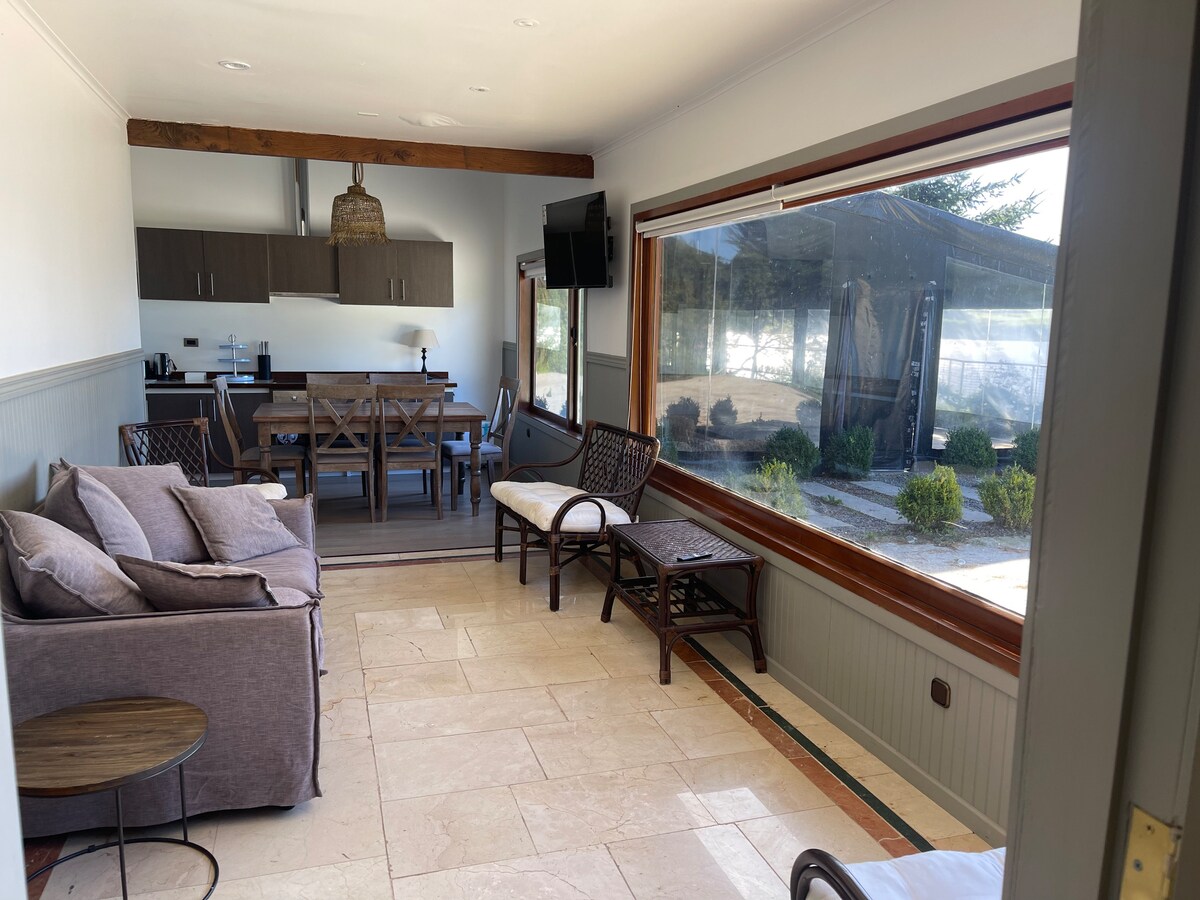 A spacious living area is visible, featuring comfortable seating arrangements, including a sofa and lounge chairs. Large windows provide abundant natural light and a view of the outdoor space. The dining area and kitchen can be seen in the background, creating an open and inviting layout.